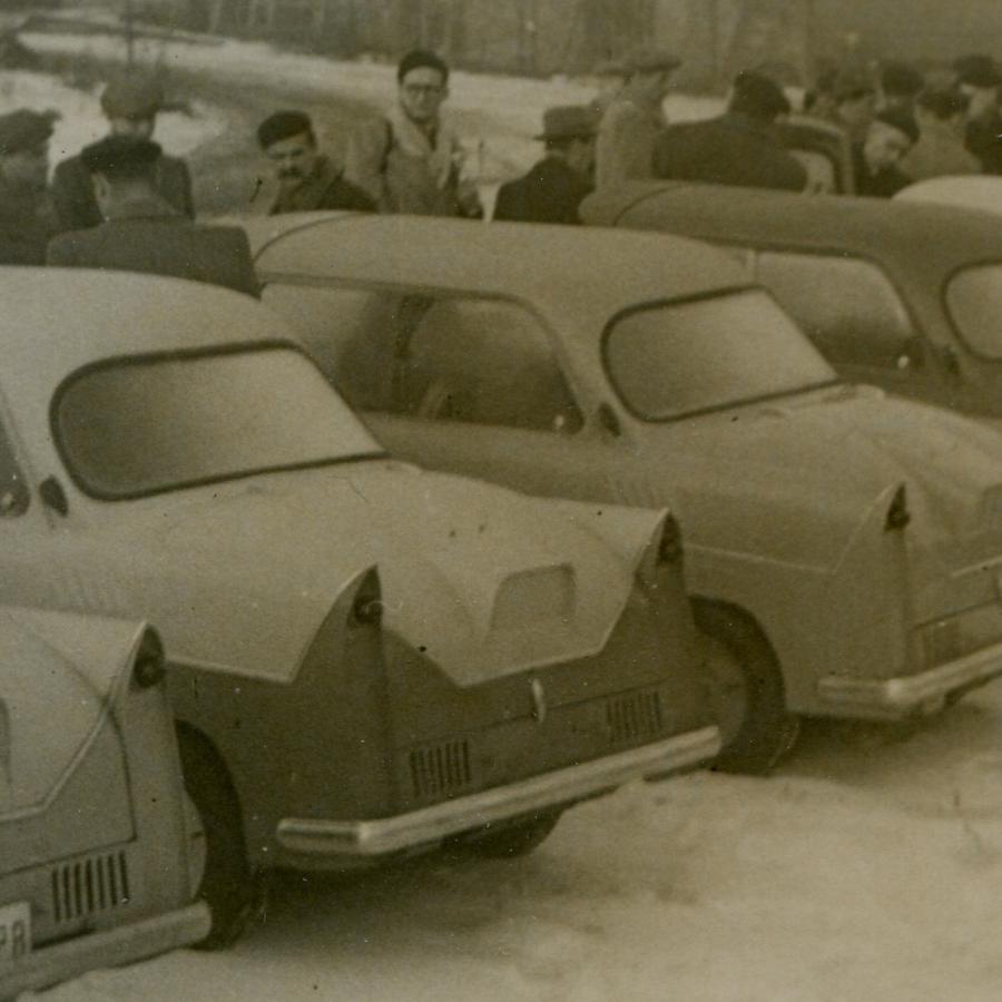 Historyczne zdjęcie w sepii. Na pierwszym planie kilka starych samochodów ustawionych tyłem, jeden obok drugiego. Za pojazdami kilkanaście osób w nieuporządkowanym rzędzie.