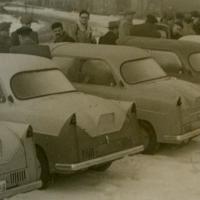 Historyczne zdjęcie w sepii. Na pierwszym planie kilka starych samochodów ustawionych tyłem, jeden obok drugiego. Za pojazdami kilkanaście osób w nieuporządkowanym rzędzie.